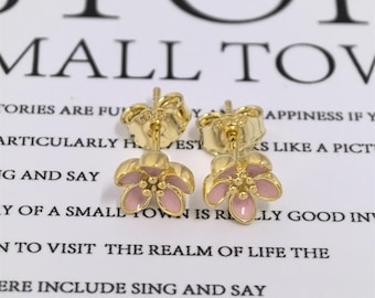 May include: A pair of gold-colored flower earrings with pink enamel petals. The earrings have a post-back closure and are set against a white background with blurred text.