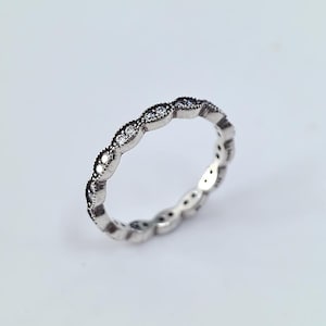 Puede incluir: Un anillo de plata con un patrón repetitivo de pequeñas piedras preciosas claras engastadas en un diseño festoneado. El anillo está sobre un fondo blanco, mostrando sus intrincados detalles y su superficie reflectante. Una delicada pieza de joyería.