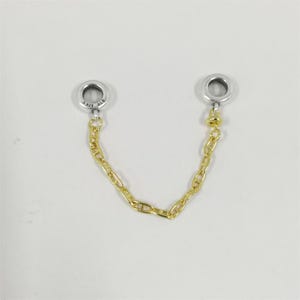 Può includere: Una catena di sicurezza per ciondoli color argento con due estremità circolari collegate da una catena color oro. Le estremità hanno un piccolo foro al centro. La catena è composta da piccole maglie.