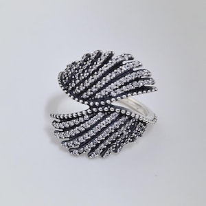 May include: A silver-toned ring with a unique design. The ring features two open, fan-like structures adorned with small, clear gemstones and dark accents. The ring's design is symmetrical and sits on a white background.