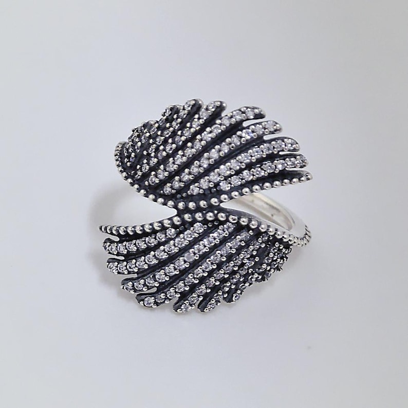May include: A silver-toned ring with a unique design. The ring features two open, fan-like structures adorned with small, clear gemstones and dark accents. The ring's design is symmetrical and sits on a white background.