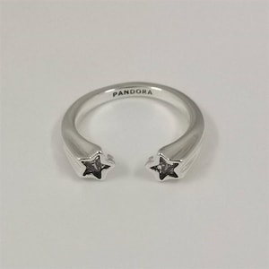 May include: A silver ring with the brand name "PANDORA" engraved on the band. The ring features two star-shaped accents, each with a dark center, at the open ends of the band. The ring has a polished finish.