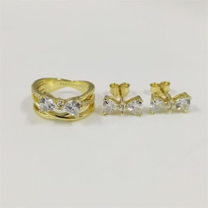 May include: A gold-toned jewelry set featuring a ring and a pair of earrings. The ring and earrings are designed with a bow shape and adorned with clear, sparkling gemstones. The ring has a double band design.