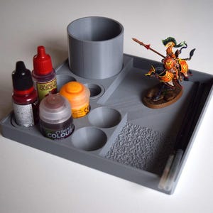 Puede incluir: Un organizador de pintura para pasatiempos de plástico gris con varios compartimentos para botes de pintura, un soporte cilíndrico y una figura en miniatura. El organizador también tiene un área texturizada y espacio para pinceles.