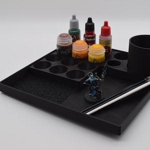 May include: A black plastic hobby organizer tray featuring a grid of circular slots for paint bottles, a larger cylindrical cup for brushes, and a flat workspace area. The tray holds several small paint bottles, two paintbrushes, and a miniature figure.