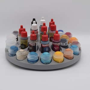 Op de afbeelding: Een cirkelvormig grijs plastic verfopbergrek met een verscheidenheid aan hobbyverfflesjes. Het rek is voorzien van meerdere verzonken ronde sleuven met druppelflesjes met rode doppen en korte potjes met verschillende gekleurde deksels, waaronder blauw, geel en wit.