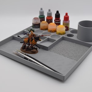 Könnte beinhalten: Eine graue Kunststoff-Malstation mit verschiedenen Fächern, darunter ein Becherhalter, Farbnäpfe und ein strukturierter Bereich. Sie enthält mehrere Farbtuben, Farbtöpfe, eine Miniaturfigur und Pinsel. Die Station ist für die Miniaturmalerei konzipiert.
