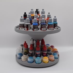 Op de afbeelding: Een tweelaags draaibaar verfrek van grijs plastic met talloze kleine druppelflesjes in diverse kleuren. De bovenste laag bevat Game Color-verf, terwijl de onderste laag een mix van verfpotjes met gekleurde deksels en etiketten bevat.