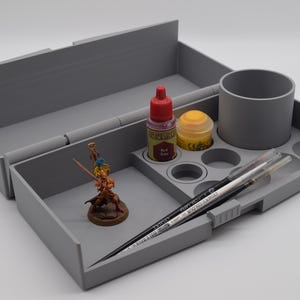 Peut inclure: Un plateau organisateur de loisirs créatifs en plastique gris avec un grand compartiment ouvert, un porte-gobelet cylindrique et plusieurs emplacements circulaires pour pots de peinture. Il contient une figurine peinte, deux pots et deux pinceaux.