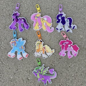 Puede incluir: Una colección de siete llaveros de personajes de dibujos animados coloridos. Los llaveros presentan varios personajes en tonos de morado, amarillo, rosa, azul y verde. Cada llavero tiene un cierre metálico.