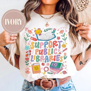 Könnte beinhalten: Cremefarbenes T-Shirt mit dem mehrfarbigen Schriftzug "SUPPORT PUBLIC LIBRARIES". Das Design umfasst Illustrationen von Büchern, Blumen und einer Rakete. Das Shirt wird mit einer blauen Jeans und einem braunen Gürtel getragen.