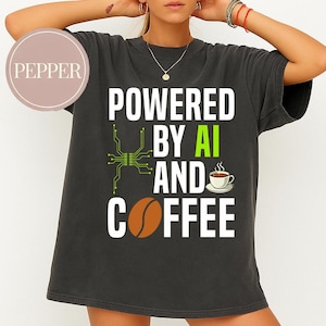 Op de afbeelding: Donkergrijs t-shirt met de tekst "POWERED BY AI AND COFFEE" in wit, groen en bruin. Het ontwerp bevat een koffiekopje, een koffieboon en een printplaat.