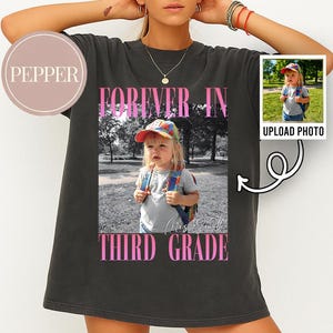 Może przedstawiać: Ciemnoszary t-shirt z niestandardowym nadrukiem graficznym. Projekt zawiera czarno-białe zdjęcie dziecka w kolorowej czapce i plecaku, z różowym napisem "FOREVER IN" powyżej i "THIRD GRADE" poniżej zdjęcia.