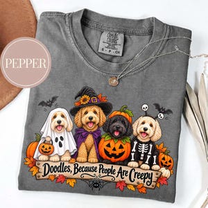 Puede incluir: Una camiseta gris carbón con un gráfico de cuatro perros goldendoodle disfrazados de Halloween, incluyendo un fantasma, una bruja, una calabaza y un esqueleto. El texto dice Doodles, Because People Are Creepy, con hojas de otoño.