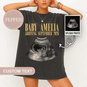May include: A charcoal gray t-shirt featuring the gold text BABY AMELIA ARRIVING SEPTEMBER 2026 above a central ultrasound image. A smaller inset box in the upper right corner displays an ultrasound scan with the text UPLOAD PHOTO.
