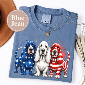 Puede incluir: Una camiseta azul de cuello redondo con un gráfico de tres perros Basset Hound. El perro de la izquierda tiene un pelaje azul con estrellas blancas, el central es blanco y el de la derecha tiene rayas rojas y blancas. Un collar dorado descansa.