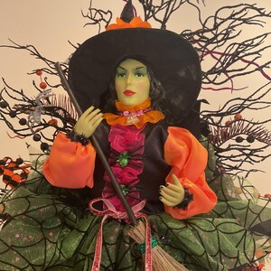 Peut inclure: Décoration de sorcière d'Halloween avec un visage vert, un chapeau noir et des accents orange. La sorcière porte une robe noire avec une jupe en toile d'araignée verte et tient un balai. Des branches décoratives et des araignées entourent la sorcière.