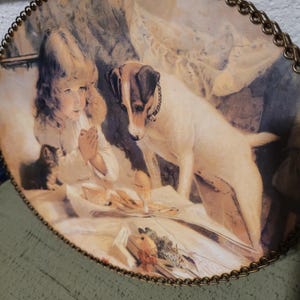 May include: Oval decorative plate featuring a vintage illustration. The image depicts a young child with a kitten and a dog, with a table set with food. The plate has a gold-colored metal trim around the edge.