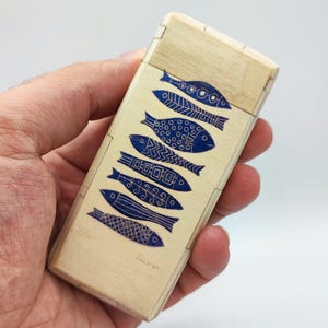 Può includere: Una scatola rettangolare di legno con coperchio incernierato. La scatola è di colore chiaro con una serie di disegni di pesci blu. I pesci variano per motivo e dimensioni. La scatola è tenuta in una mano.
