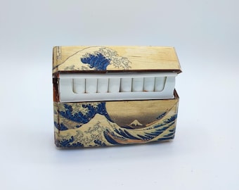 May include: A wooden cigarette case featuring a print of Hokusai's "The Great Wave off Kanagawa." The case is rectangular with a hinged lid, and the interior holds a row of cigarettes. The artwork depicts a large blue wave and a mountain.