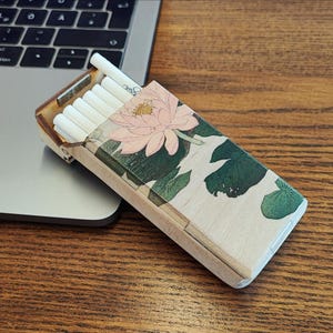 Puede incluir: Un estuche de cigarrillos de madera con un diseño floral que presenta una flor de loto rosa y hojas de nenúfar verdes. El estuche está abierto, revelando varios cigarrillos. El estuche está sobre una superficie de madera junto a un portátil.