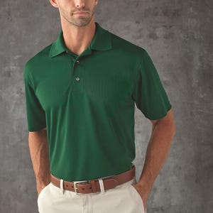 Puede incluir: Un polo verde con cuello y mangas cortas. La camisa se combina con pantalones color caqui y un cinturón de cuero marrón con hebilla plateada. El modelo está de pie frente a un fondo gris.