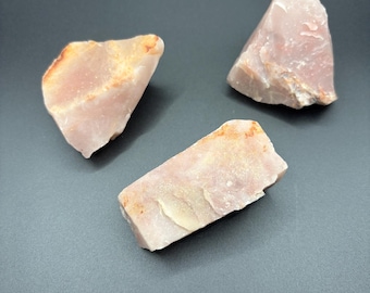 Puede incluir: Tres piedras en bruto y sin pulir con una forma irregular y dentada. Las piedras son de un color rosa pálido y polvoriento con sutiles parches de depósitos minerales de color crema y naranja claro en sus superficies irregulares.