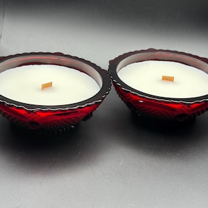 May include: Two red glass bowl candles with white wax and wooden wicks. The bowls have a decorative, textured design and a dark rim. The candles are set against a dark background.