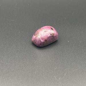 Puede incluir: Una piedra preciosa pulida y rodada con un tono magenta vibrante y rosa intenso. La piedra presenta una superficie lisa y redondeada con inclusiones naturales visibles y vetas minerales oscuras e irregulares en su estructura.