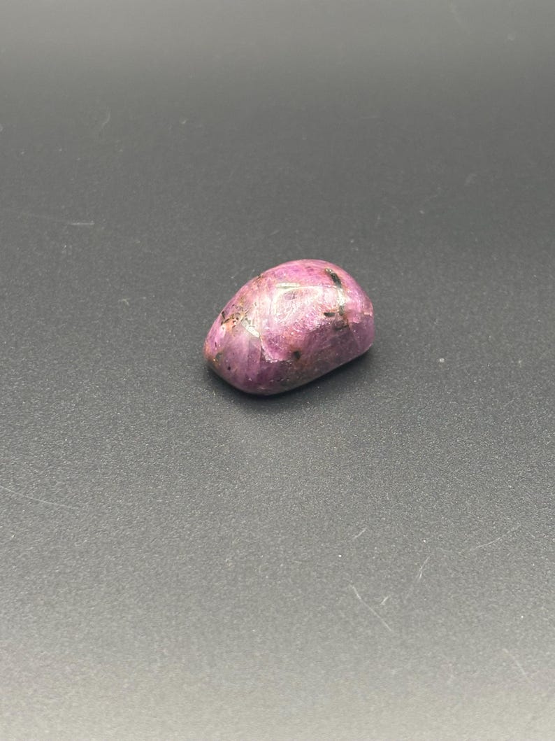 Puede incluir: Una piedra preciosa pulida y rodada con un tono magenta vibrante y rosa intenso. La piedra presenta una superficie lisa y redondeada con inclusiones naturales visibles y vetas minerales oscuras e irregulares en su estructura.