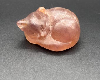 Puede incluir: Una pequeña figura tallada de un gato durmiendo, hecha de una piedra translúcida de color rosa polvoriento. La escultura presenta contornos suaves y redondeados que definen la postura enroscada del felino, con inclusiones minerales internas visibles y un acabado pulido.