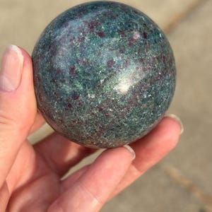 May include: A polished spherical stone featuring a deep green base color with scattered, vibrant magenta and crimson inclusions throughout the surface. The mineral sphere displays a smooth, reflective texture with natural variations in color and pattern.
