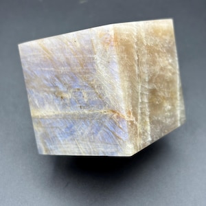 May include: A polished, cube-shaped moonstone with a mix of beige, cream, and pale blue hues. The stone has a subtle, iridescent sheen and a smooth, tactile surface. The cube is displayed against a dark gray background.