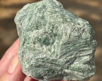 Puede incluir: Un trozo bruto y sin pulir de mineral verde con una textura fibrosa y estriada. La piedra muestra varios tonos de verde bosque y salvia con una superficie cristalina ligeramente reflectante y bordes irregulares y dentados.