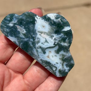 May include: A polished moss agate stone carved into a cloud shape, featuring a smooth surface with intricate, swirling patterns of deep forest green inclusions set against a translucent, milky white and clear crystalline background.