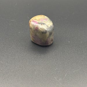 Puede incluir: Una piedra preciosa pulida y rodada que presenta una superficie lisa y redondeada con un aspecto moteado. La piedra muestra una mezcla de tonos opacos crema, beige y gris, acentuados por parches distintos e irregulares de magenta y rosa.