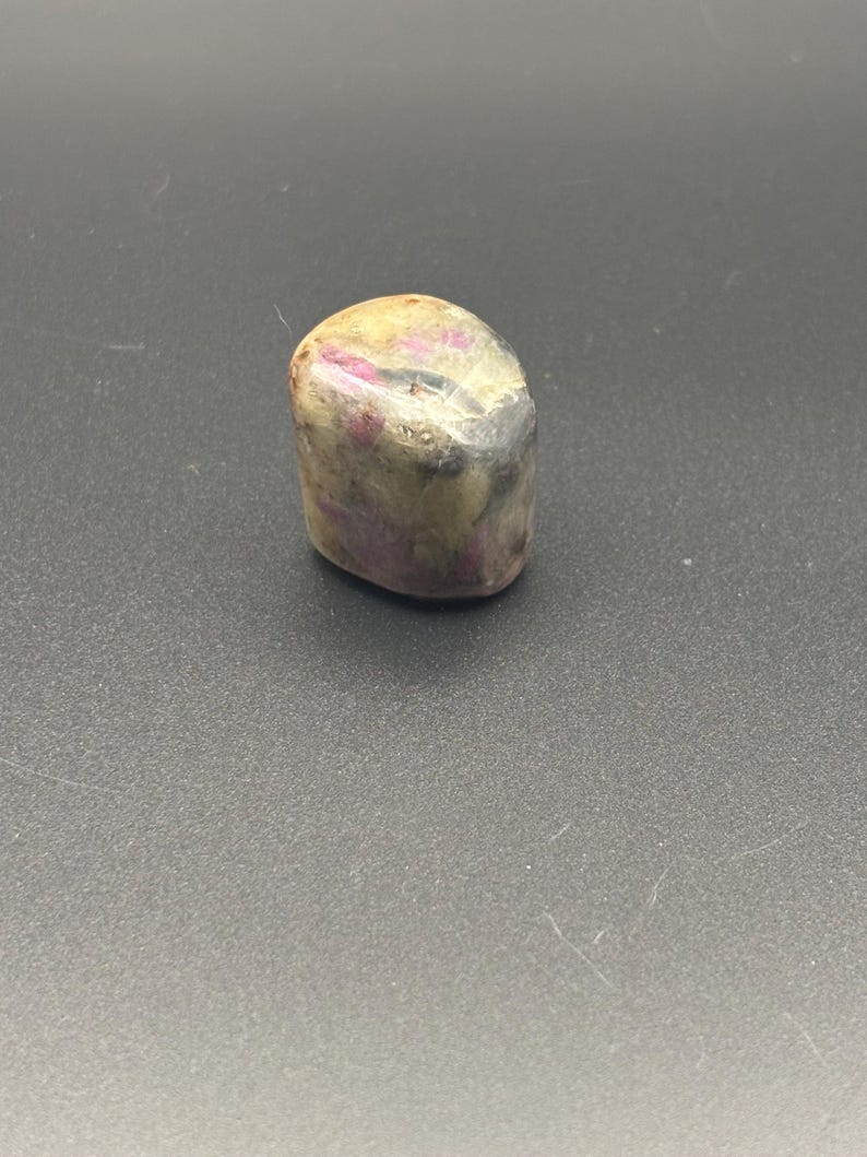 Puede incluir: Una piedra preciosa pulida y rodada que presenta una superficie lisa y redondeada con un aspecto moteado. La piedra muestra una mezcla de tonos opacos crema, beige y gris, acentuados por parches distintos e irregulares de magenta y rosa.