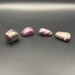May include: Four polished tumbled stones arranged in a row. Each stone features a unique combination of deep magenta, pink, and greyish-white mineral inclusions. The stones have smooth, rounded surfaces and irregular shapes with varying color patterns.