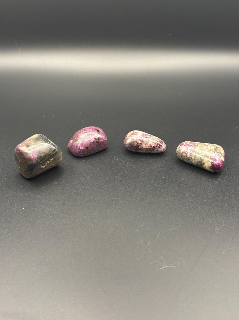 Puede incluir: Cuatro piedras pulidas dispuestas en fila. Cada piedra presenta una combinaci&oacute;n &uacute;nica de inclusiones minerales en color magenta intenso, rosa y blanco gris&aacute;ceo. Las piedras tienen superficies lisas y redondeadas con formas irregulares.