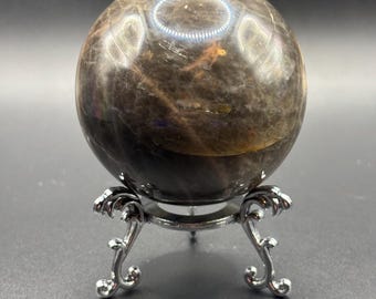 Puede incluir: Una piedra esférica pulida con un aspecto jaspeado en marrón oscuro y negro, que presenta sutiles inclusiones doradas y una superficie reflectante. La esfera descansa de forma segura sobre un soporte de metal plateado con tres patas curvas y ornamentadas.