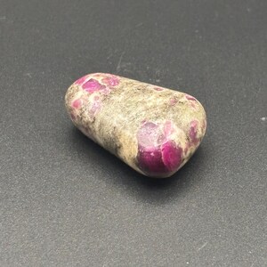 Puede incluir: Una piedra pulida y rodada que presenta una forma lisa y redondeada con una superficie moteada. La piedra muestra una combinaci&oacute;n de tonos tierra opacos en gris claro y beige, intercalados con inclusiones cristalinas de color magenta intenso.