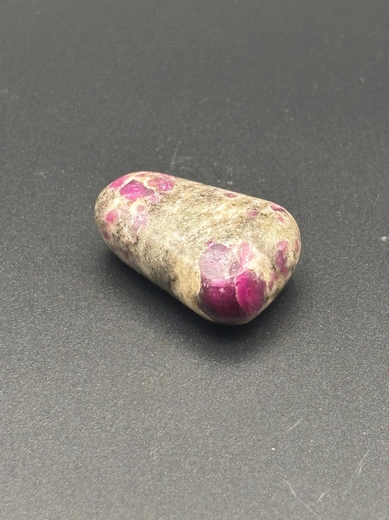 Puede incluir: Una piedra pulida y rodada que presenta una forma lisa y redondeada con una superficie moteada. La piedra muestra una combinaci&oacute;n de tonos tierra opacos en gris claro y beige, intercalados con inclusiones cristalinas de color magenta intenso.