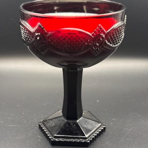 May include: A deep red glass goblet featuring a textured diamond pattern around the bowl. The stem and the hexagonal base are made of solid black glass, providing a stark contrast to the vibrant red upper portion of the drinking vessel.