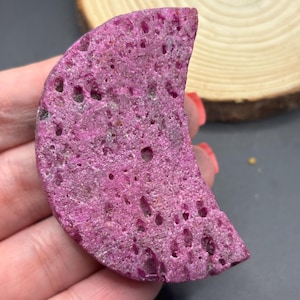 May include: A crescent moon-shaped piece of raw, vibrant magenta-colored mineral with a porous, textured surface. The stone features numerous small, irregular indentations and a rough, crystalline appearance throughout its entire structure.