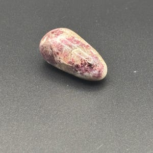 Puede incluir: Una piedra pulida y lisa con una forma irregular y alargada. La superficie presenta un patr&oacute;n moteado de tonos magenta intenso y rosa mezclados con secciones de color blanco crema y cristalino transl&uacute;cido, creando una textura variada.