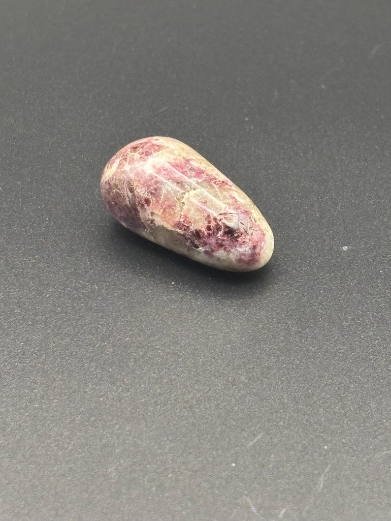 Puede incluir: Una piedra pulida y lisa con una forma irregular y alargada. La superficie presenta un patr&oacute;n moteado de tonos magenta intenso y rosa mezclados con secciones de color blanco crema y cristalino transl&uacute;cido, creando una textura variada.