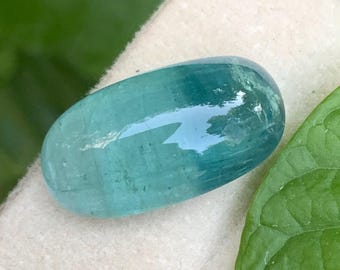 May include: A smooth, oval-shaped gemstone with a translucent, teal-blue color and a polished, reflective surface. The stone displays subtle internal variations and a glassy luster, showcasing a vibrant, cool-toned appearance throughout its body.