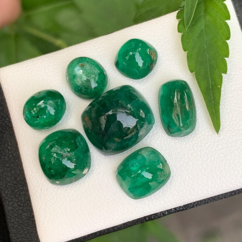 May include: A collection of seven loose, polished emerald gemstones in various shapes, including round, oval, and cushion cuts. The stones exhibit a deep, vibrant green color with natural inclusions and a smooth, reflective surface finish.