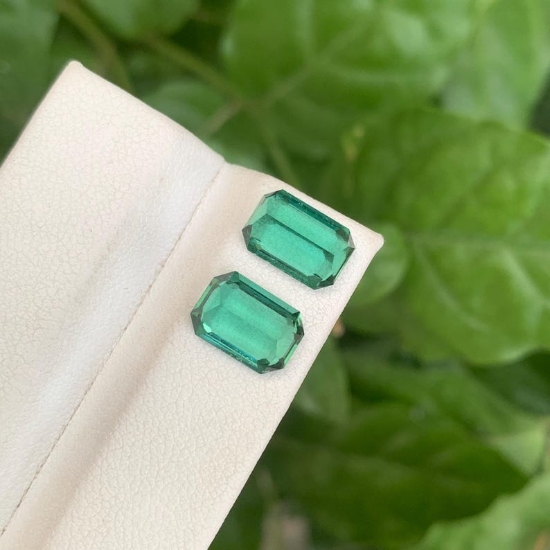 May include: Two emerald-cut, vibrant green gemstones. The gems are displayed on a white surface, with a blurred green background. The gemstones have a rectangular shape with beveled edges, reflecting light. The image showcases the gemstones' clarity and color.