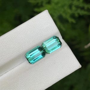 May include: Two rectangular, teal-colored gemstones with a bright, reflective surface. The gems are set on a white surface, with a blurred green background. The gems have a rectangular cut with visible facets.